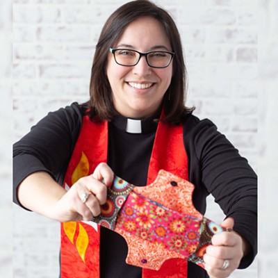 A woman with shoulder-length brown hair and glasses in a pastor's frock and color holding a red patterned fabric maxi pad