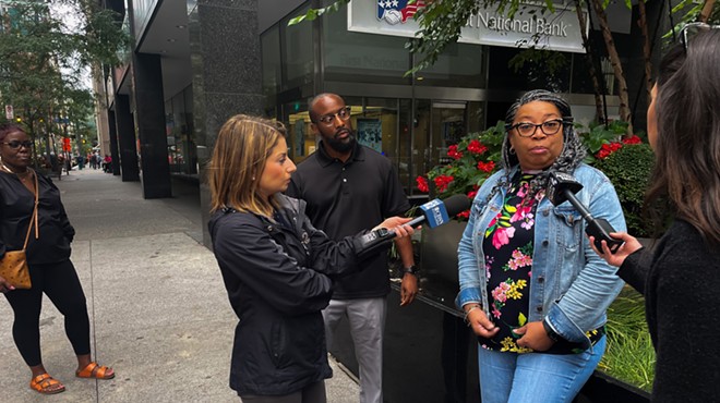 A woman in glasses in a denim jacket and pants speaks with two television reporters holding microphones