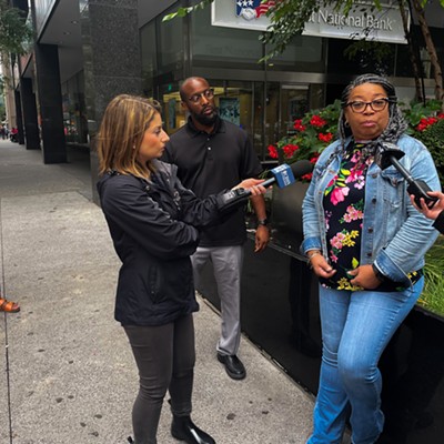 A woman in glasses in a denim jacket and pants speaks with two television reporters holding microphones