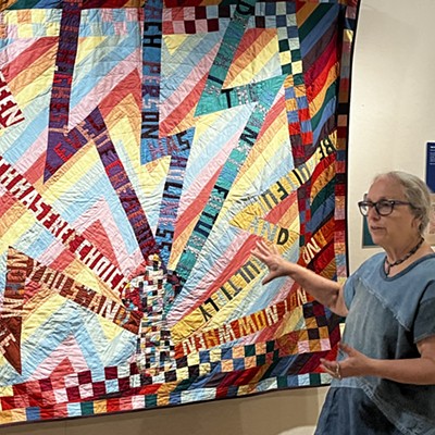 A woman in a collaged denim dress and glasses stands near a quilt of neon lightning bolts