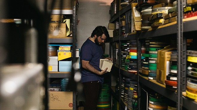 A dark-haired bearded man looks through film reels and boxes of film supplies on shelves in a basement