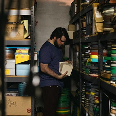 A dark-haired bearded man looks through film reels and boxes of film supplies on shelves in a basement