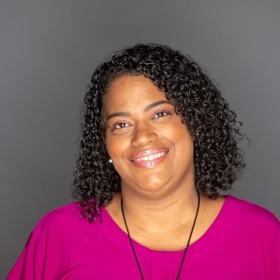 A woman with brown skin and tightly curled hair down to her shoulders wears a pink shirt and necklace