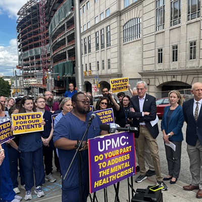 A nurse with short hair, a beard and glasses speaks before a crowd in scrubs and suits near large hospital buildings