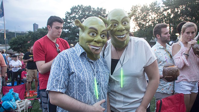 Two concert attendees wear Shrek masks and glo-stick necklaces, with one giving the thumbs up.