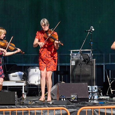 Five people on a stage playing violins, a guitar, and a bass in front of a sign that says "Irish Festival Est. 1991"