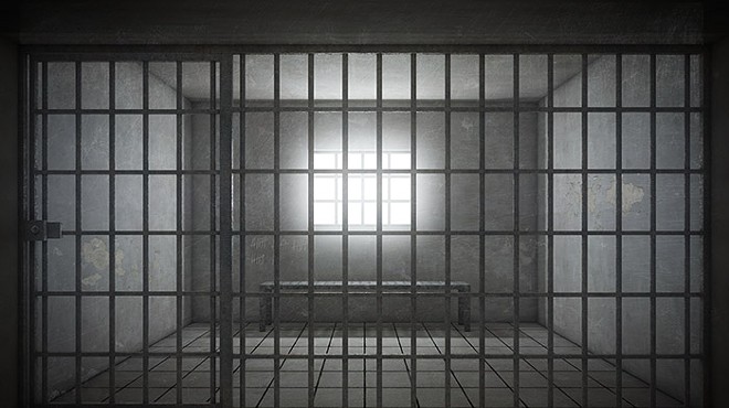 Empty prison cell showing bars, an empty bench, and light coming in through the window