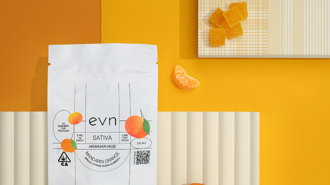 White bag of THC/CBD gummies with three oranges on the label, text reads: "evn SATIVA HAWAIIN HAZE MANDARIN ORANGE single strain rosin gummies". Abstract yellow and orange background with a mandarin orange and a little pile of orange gummies  in the frame.