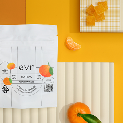 White bag of THC/CBD gummies with three oranges on the label, text reads: "evn SATIVA HAWAIIN HAZE MANDARIN ORANGE single strain rosin gummies". Abstract yellow and orange background with a mandarin orange and a little pile of orange gummies  in the frame.
