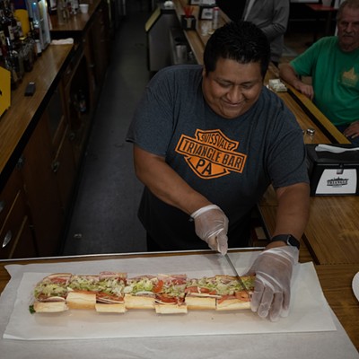 A man smiles as he cuts a two-foot hoagie into three-inch sections on a wooden bar