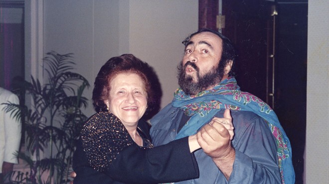 A short aging woman with frizzy dark hair dances with a large man pantomiming opera singing