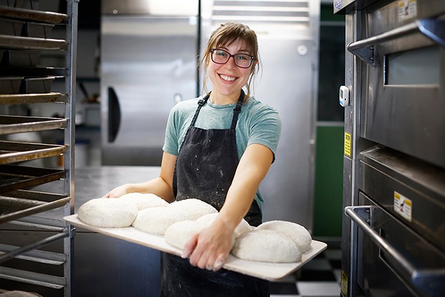 A woman wearing an apron holds a tray of bread dough in a bakery kitchen