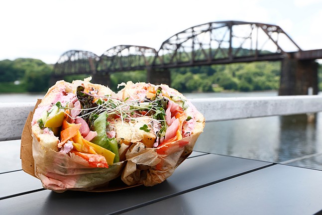 A large veggie sandwich sits on a table, with a river and bridge in the background