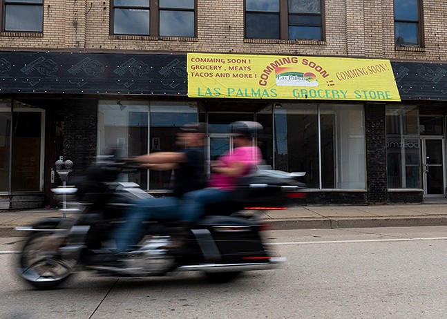 A fast-speeding motorcycle passes by the front of a building with a bright sign that says "Coming Soon!! Las Palmas Grocery Store"
