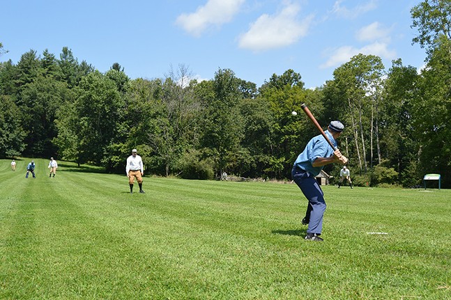 A person up to bat waiting on a pitcher during a baseball game. Other men are in the outfield. The game is being held on bright green grass, with green trees in the background, under a bright blue sky