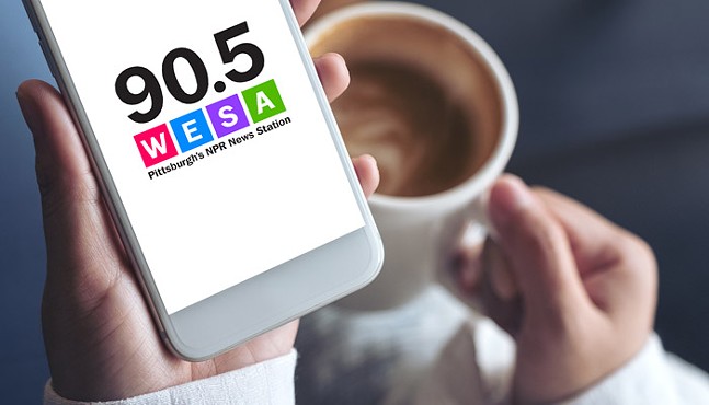 A person holding a cup of coffee in one hand and a cell phone in the other with 90.5 WESA's logo on the screen of the phone