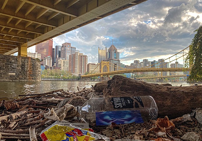 The city of Pittsburgh is foregrounded by a pile of trash photographed under a bridge.