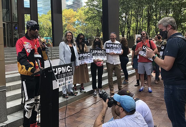 On the left, a Black man wearing sunglasses stands at a podium in front of the steps of the Steel Building. He is addressing supporters at a rally and standing next to local politicians and organizers.