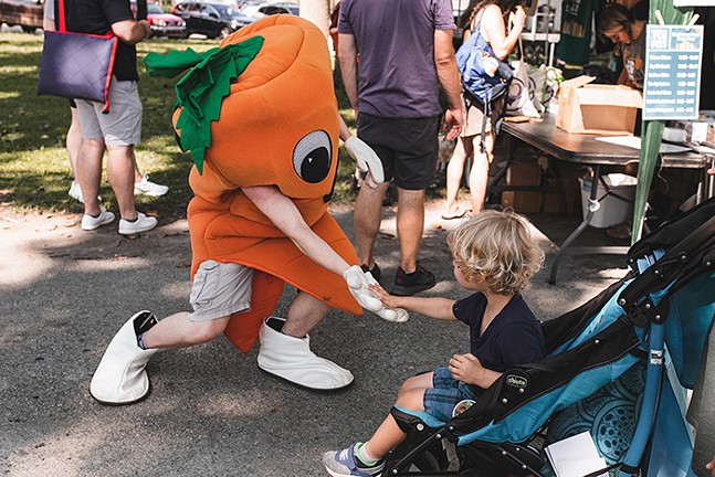A person dressed up as a carrot high fives a young child in a stroller