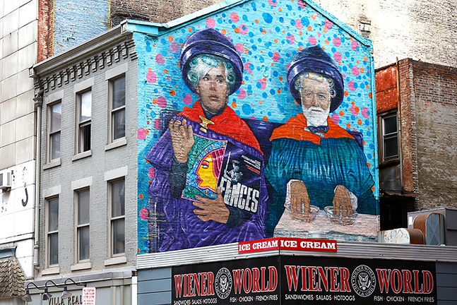 A brightly colored mural of two men sitting side by side under hair dryers at a beauty salon sits on the side of a building above a smaller building that says "Wiener World"