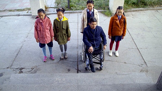 Film still of people, including one in a wheelchair, looks at a set of stairs with no ramp.
