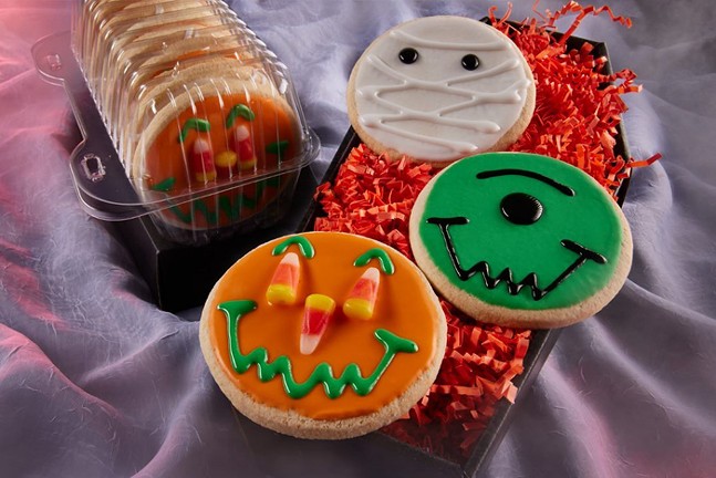 Halloween-themed donuts. One has a pumpkin face made from orange and green icing and candy corn. Another has green and black icing drawn to create a one-eyed monster face. The other looks like a mummy with white icing and black eyes