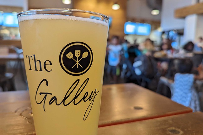 A glass of beer that says "The Galley" sitting on a table with people in the background of a dining room