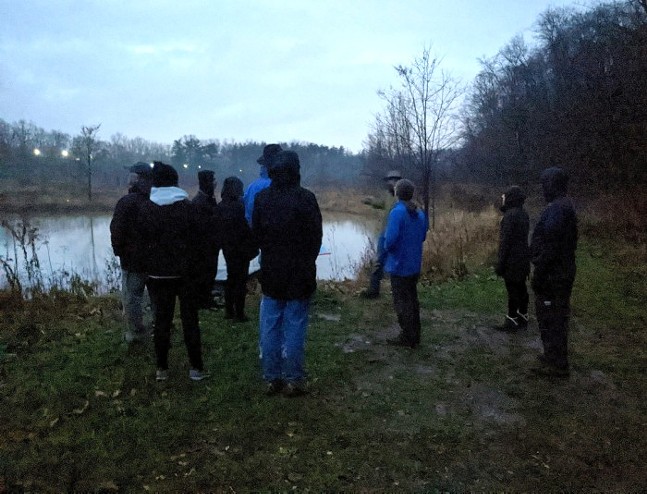 A small crowd of people in fall jackets and hoodies stands around a lake while a guide points