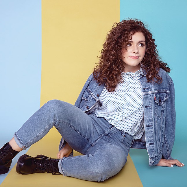 Young woman with curly dark hair, wearing jeans and a jean jacket and black boots, sits on the floor in front of a striped backdrop of light blue, yellow, and bright blue.