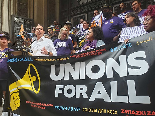 A man in a white shirt and glasses, backed by a large crowd of people, talks into a microphone in front of a huge black vinyl sign with an illustration of a yellow megaphone beside text that says "Unions for All" in multiple languages.