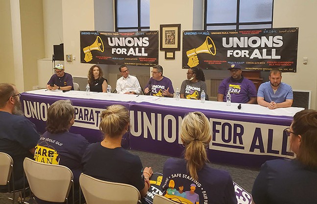People sit on chairs watching a row of seven panelists sitting at a table. Multiple signs that say "Unions for All" hang on the table and wall behind the panelists.