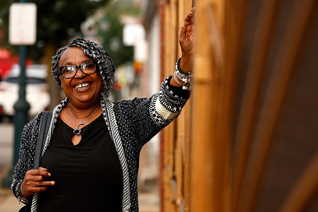 A smiling Black woman with dark-and-light gray braids stands outside holding her hand against a wall.