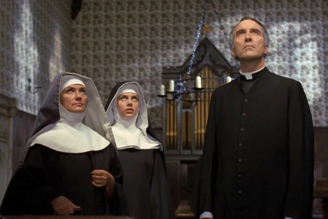 Actor Christopher Lee wears a priest outfit and stands next to two nuns in the film To the Devil a Daughter.