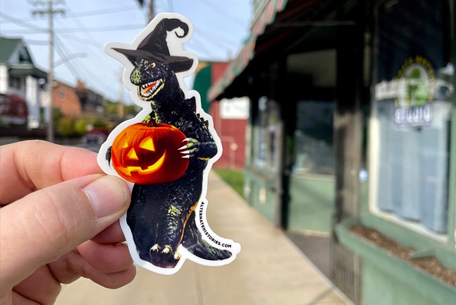 A hand holds a sticker of Godzilla wearing a witch hat and holding a jack-o-lantern.