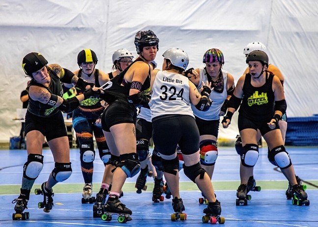 Female roller derby skaters wearing helmets, padding, tank tops, and shorts slam into each other during a competition.