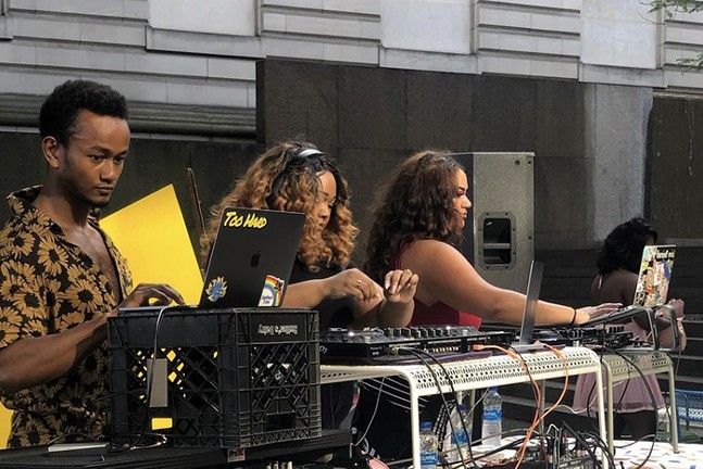 Three Black DJs stand lined up behind a table with laptops, turntables, and other equipment.