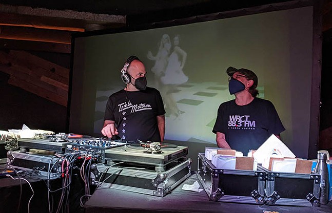 Two DJs wearing face masks glance at each other as a projection of female dancers plays in the background.