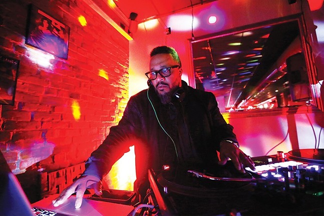 A DJ reaches over a table as multicolored lights shine behind him.