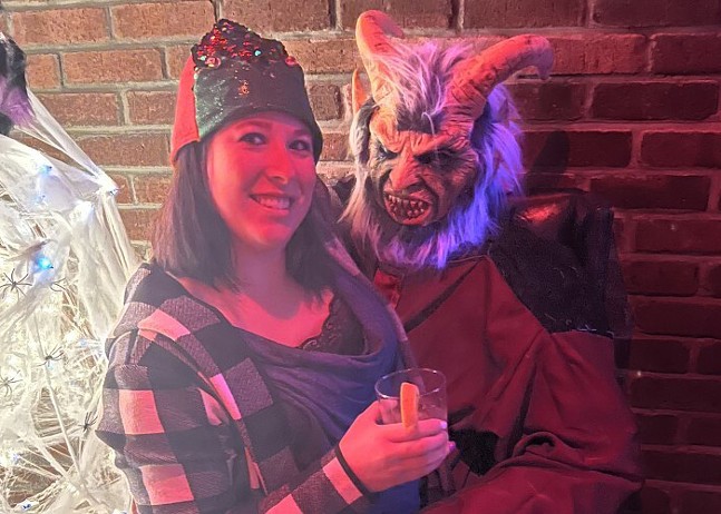 A woman smiles at the camera as she sits on the lap of a person dressed as Krampus