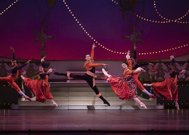 A group of ballet performers in vivid red costumes jump and spin on stage.