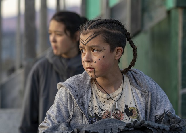 A young Inuit girl with braids and face paint looks off into the distance.