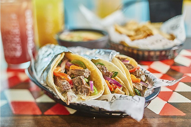 Steak tacos in flour tortillas rest in a food basket on a table filled with various Mexican cuisine.