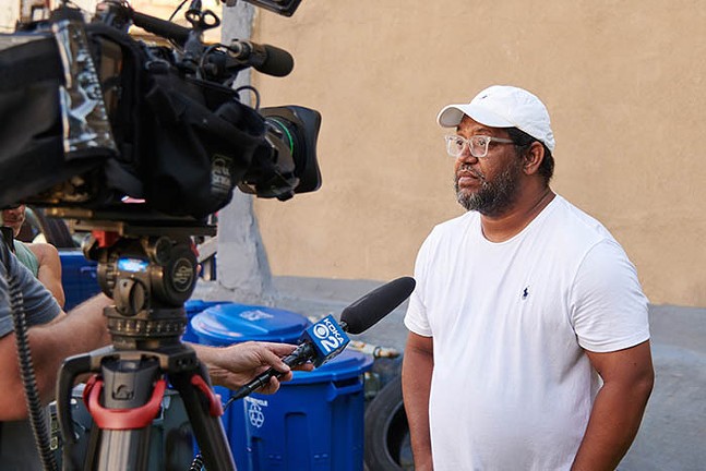 A Black man in a white T-shirt and hat is interviewed by a news crew.