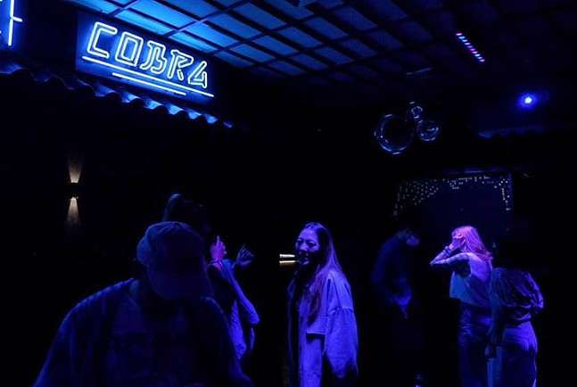 A crowd stands under a light blue neon light that spells "Cobra."