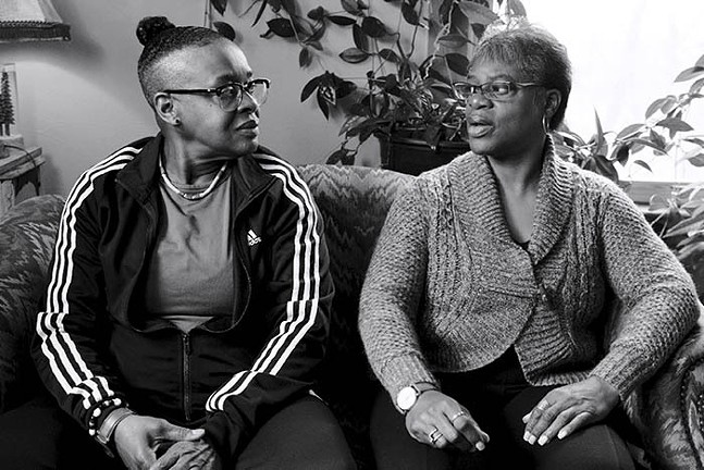 A black and white image shows two older Black women, one wearing a track jacket and glasses, the other wearing a cardigan and glasses, seated on a couch talking.