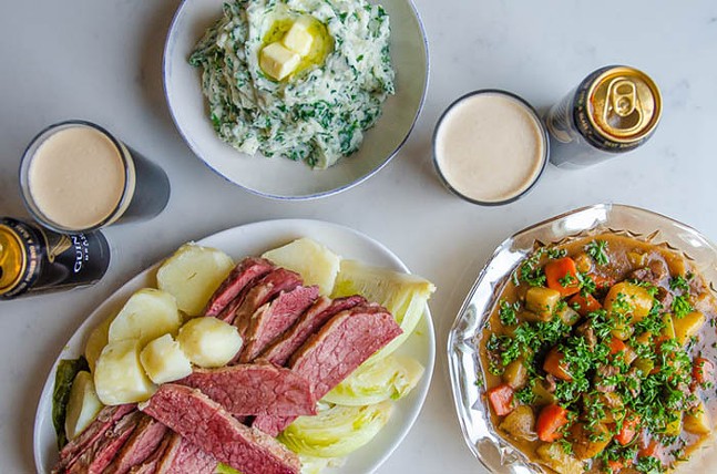 An overhead shot shows a variety of traditional Irish food, including plated corned beef, bright green and white Colcannon mashed potatoes, stew, and Guinness beer poured in a glass.