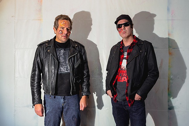 Two older white men wearing black leather jackets and jeans pose against a white wall while wearing zombie prosthetics on their faces.