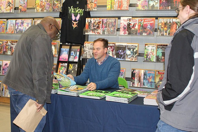 A white man seated speaks with a Black man as he leans in to look at a table full of bright green Hulk comic books. Rows of comic books and a Venom T-shirt are displayed in the background.