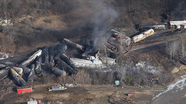 Cylindrical train cars all piled up along a railway, blackened with smoke billowing up from the wreck