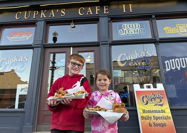 Two kids, one a boy with red hair and glasses, the other a little girl with brown hair and a pink tie-dyed outfit, hold plates of fish and chips while standing in front of Cupka's Cafe.